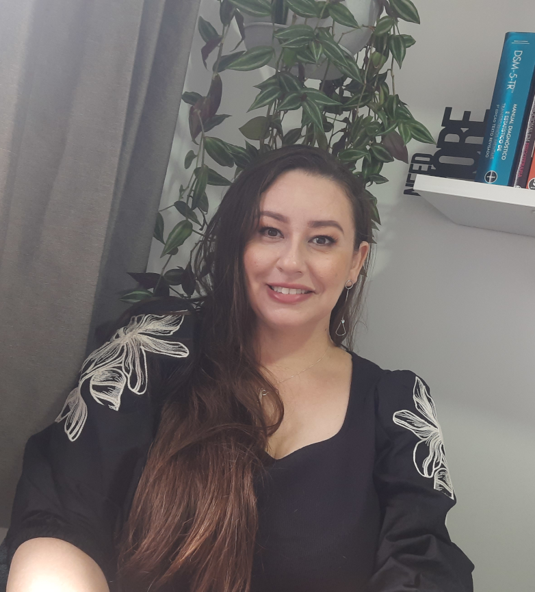 Psicóloga Fernanda olhando para camera com expressão descontraida, com planta e prateleira com livros de psicologia ao fundo