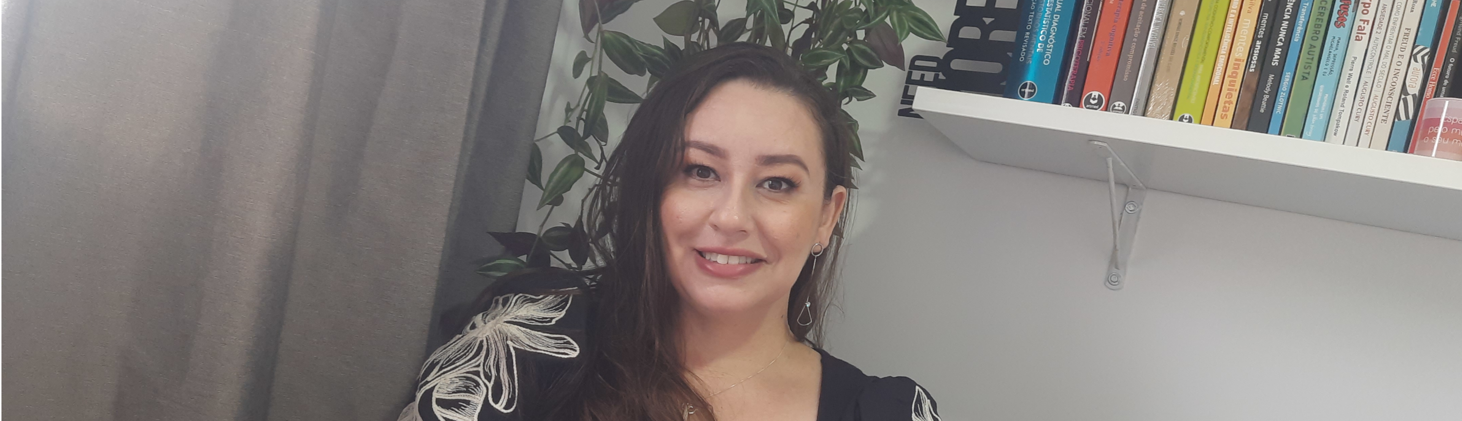 Psicóloga Fernanda olhando para camera com expressão descontraida, com planta e prateleira com livros de psicologia ao fundo