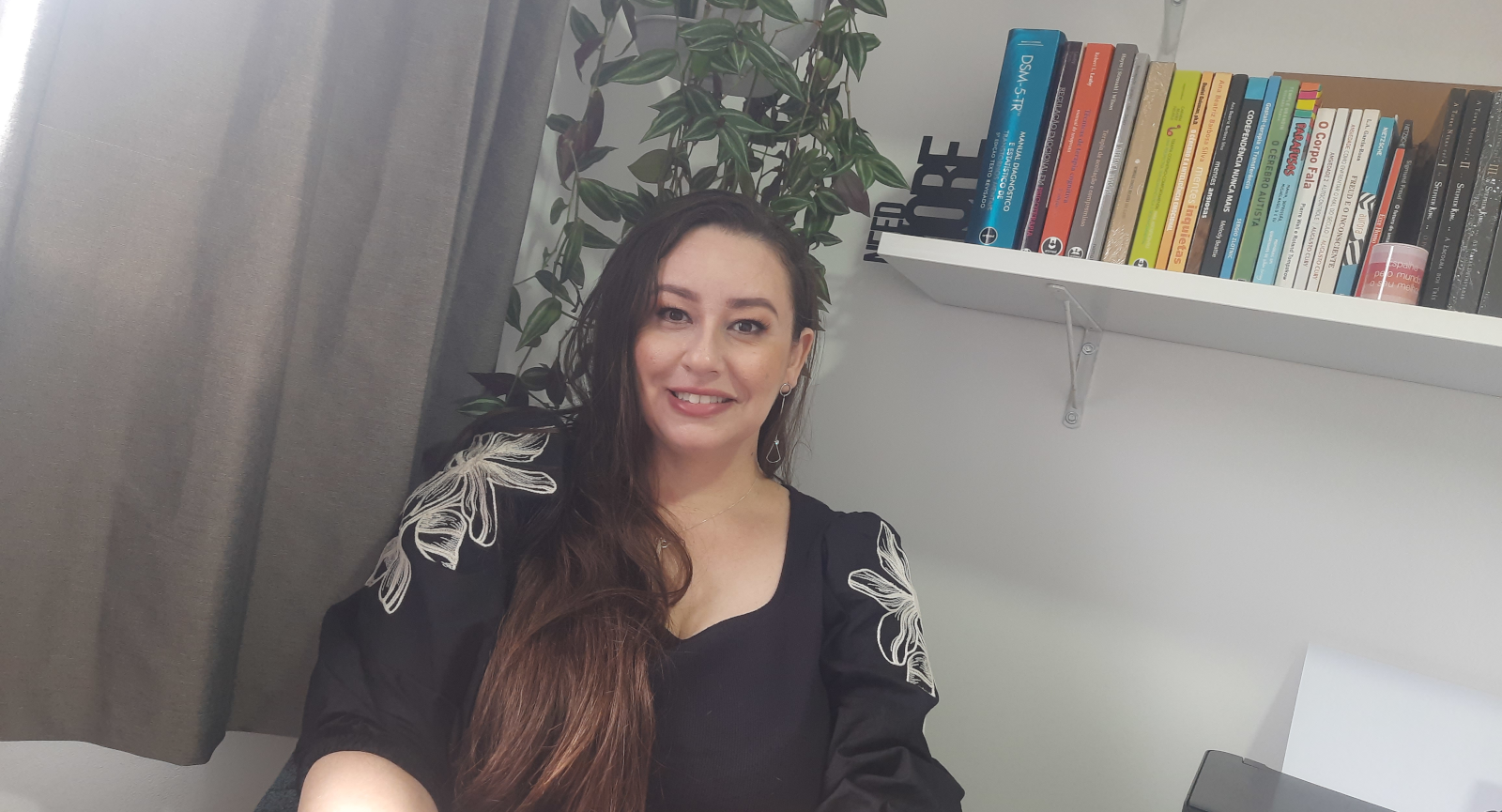 Psicóloga Fernanda olhando para camera com expressão descontraida, com planta e prateleira com livros de psicologia ao fundo